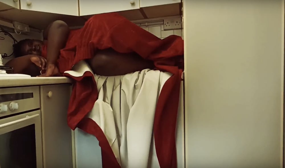 A Black person draped in red curtain lying on kitchen counter.